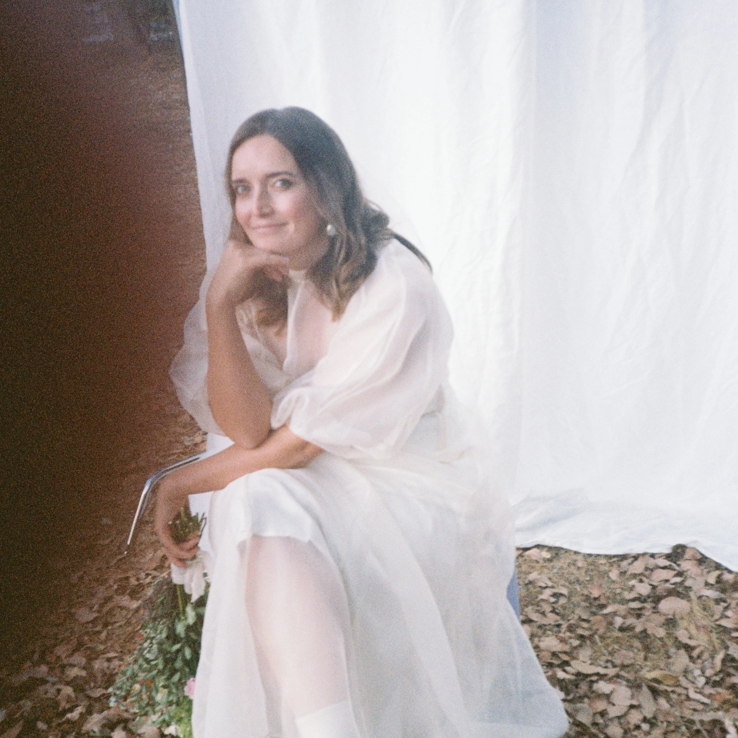 A woman in a white dress sitting on the ground with leaves, holding flowers, and smiling at the camera.