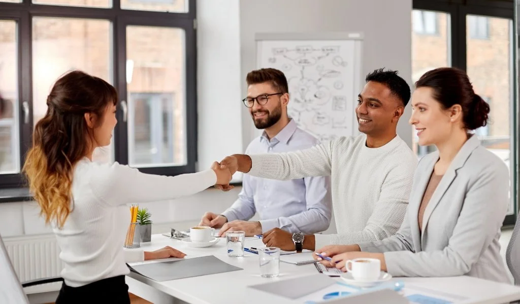A woman shaking hands with a man across a conference table, while two other people look on and smile. The setting is a modern office with large windows and a whiteboard in the background.