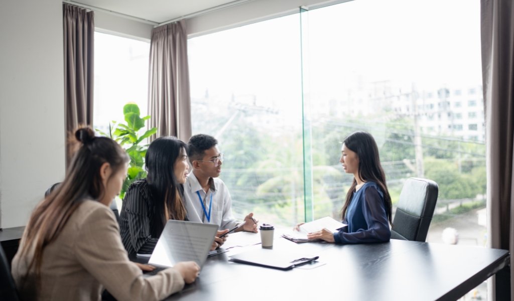 Four people sitting around a conference table in a modern office, engaged in a discussion with a woman seated at the head of the table. Large windows in the background reveal a cityscape with trees and buildings.