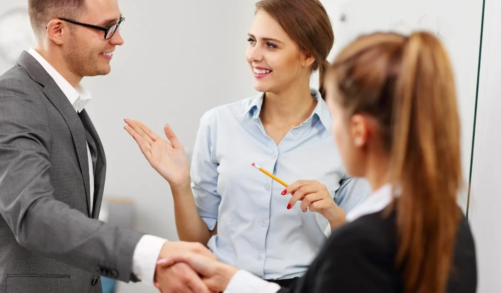 A man in a gray suit shaking hands with a woman in a black blazer; another woman with a pencil and light blue shirt is smiling and gesturing as they engage in a conversation.