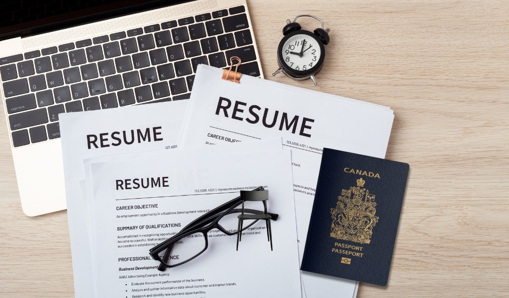 A workspace with a laptop, two resumes, a pair of glasses, a Canadian passport, a small black clock, and a miniature black chair on a wooden surface.
