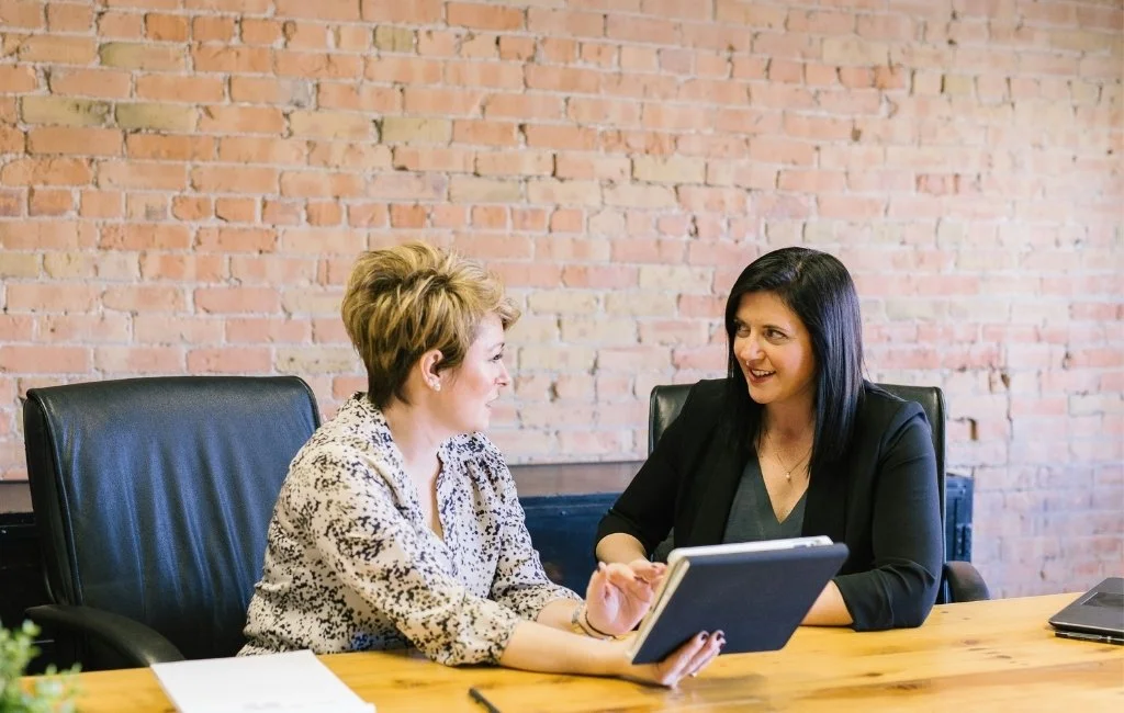Two women are sitting at a wooden table in a meeting room with a brick wall in the background. They are engaged in conversation, with one holding a tablet and the other listening attentively.