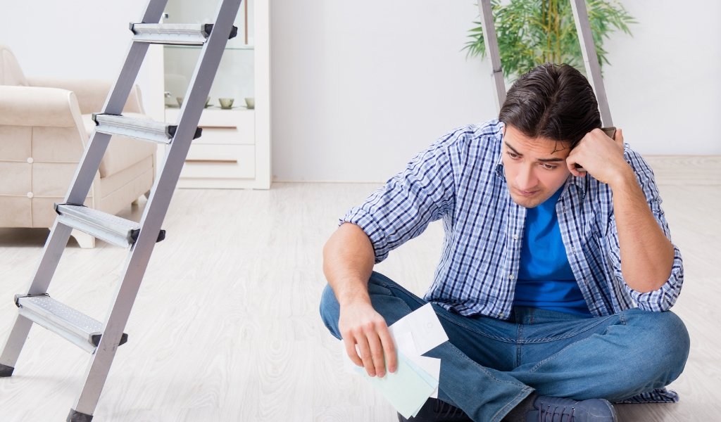 Man sitting on the floor looking worried, holding paperwork, next to a ladder, in a living room with white walls and a plant.
