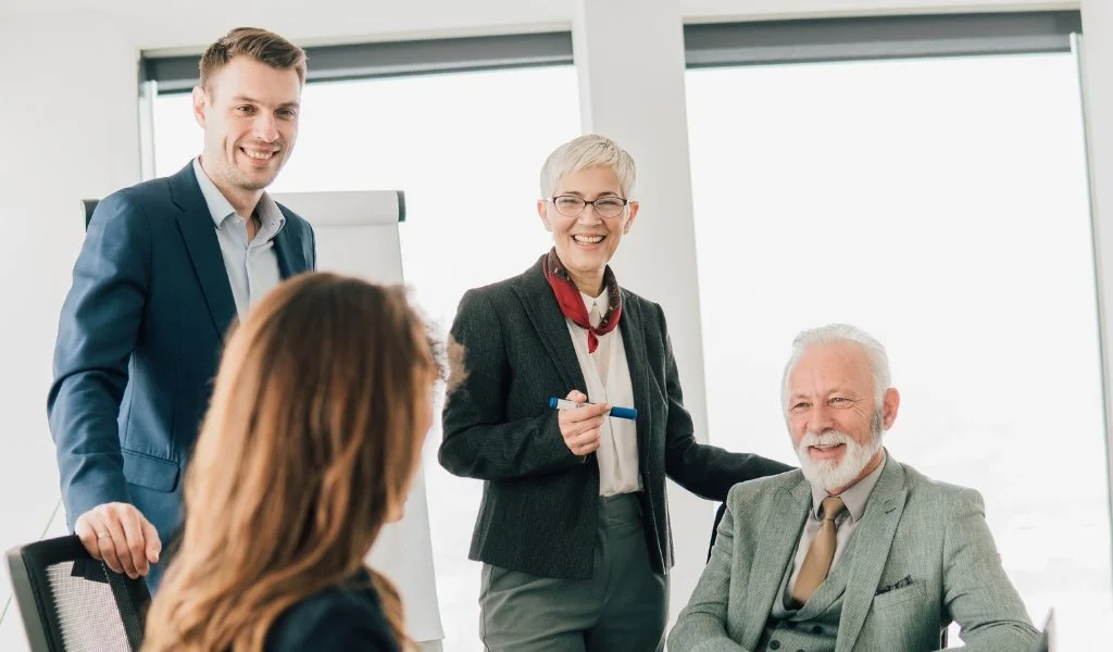Four business professionals in a bright office, with two women and two men, engaged in a discussion. One woman is standing with a marker in her hand, smiling and wearing glasses and a blazer, while an older man is sitting, also smiling, and dressed in a suit. The other two individuals are seated or standing nearby, listening attentively.