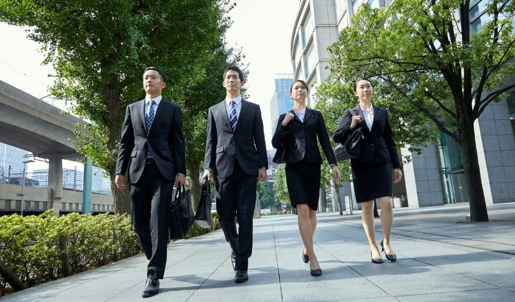 Four business professionals, two men and two women, walking on a city sidewalk in suits carrying briefcases and handbags.