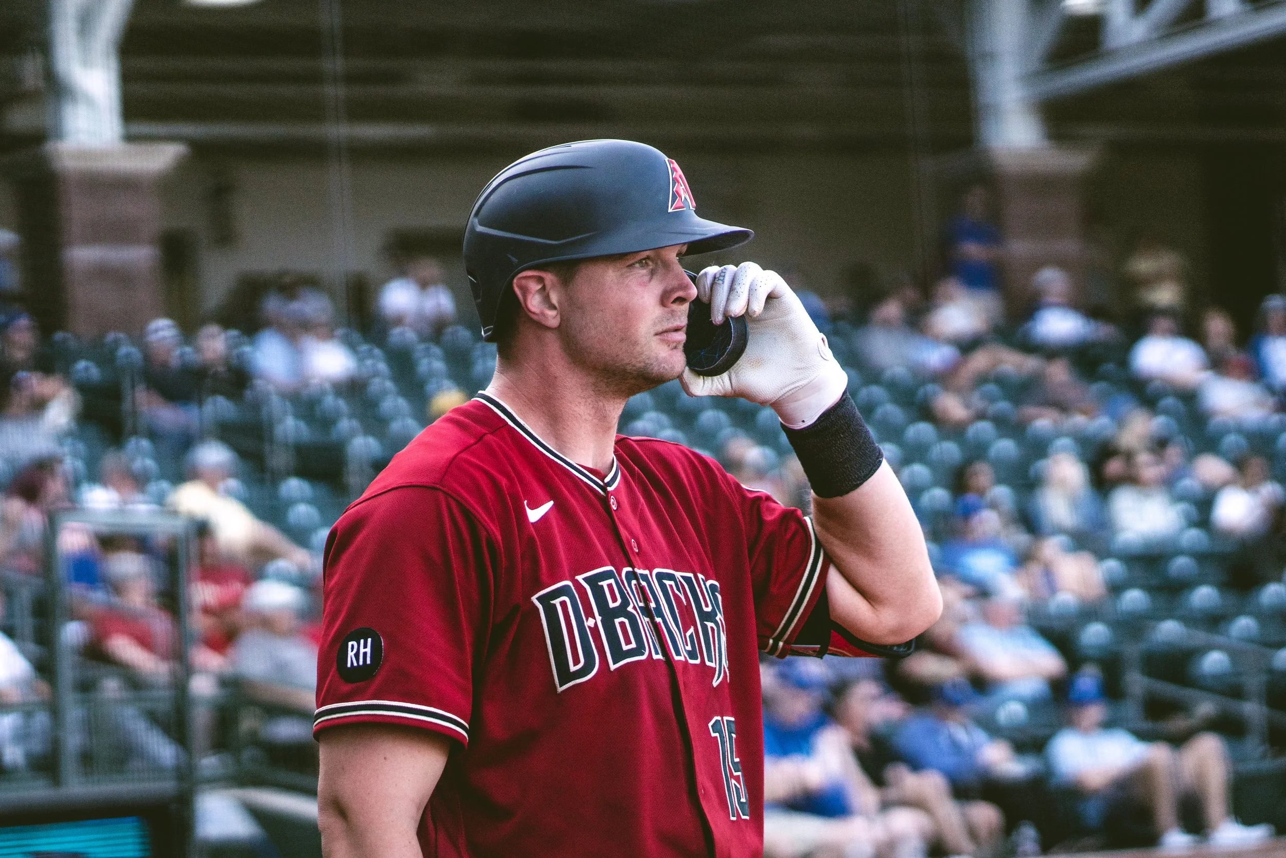 Baseball player wearing a red D-backs jersey and helmet, on the phone at a stadium with spectators in the background.