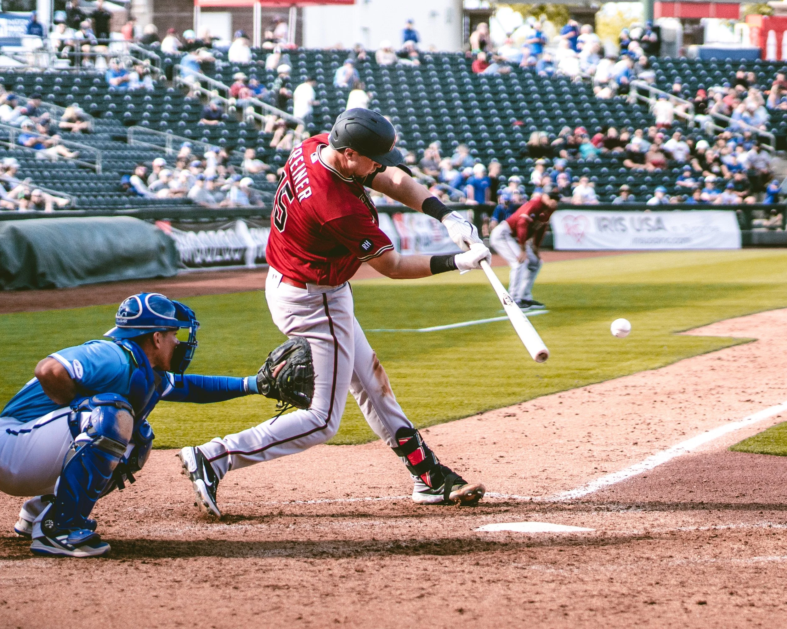 A baseball player in a red jersey and white pants is swinging a bat at a pitched ball near home plate. A catcher in blue protective gear is crouching behind him, and an umpire is behind the catcher. The stands are filled with spectators.