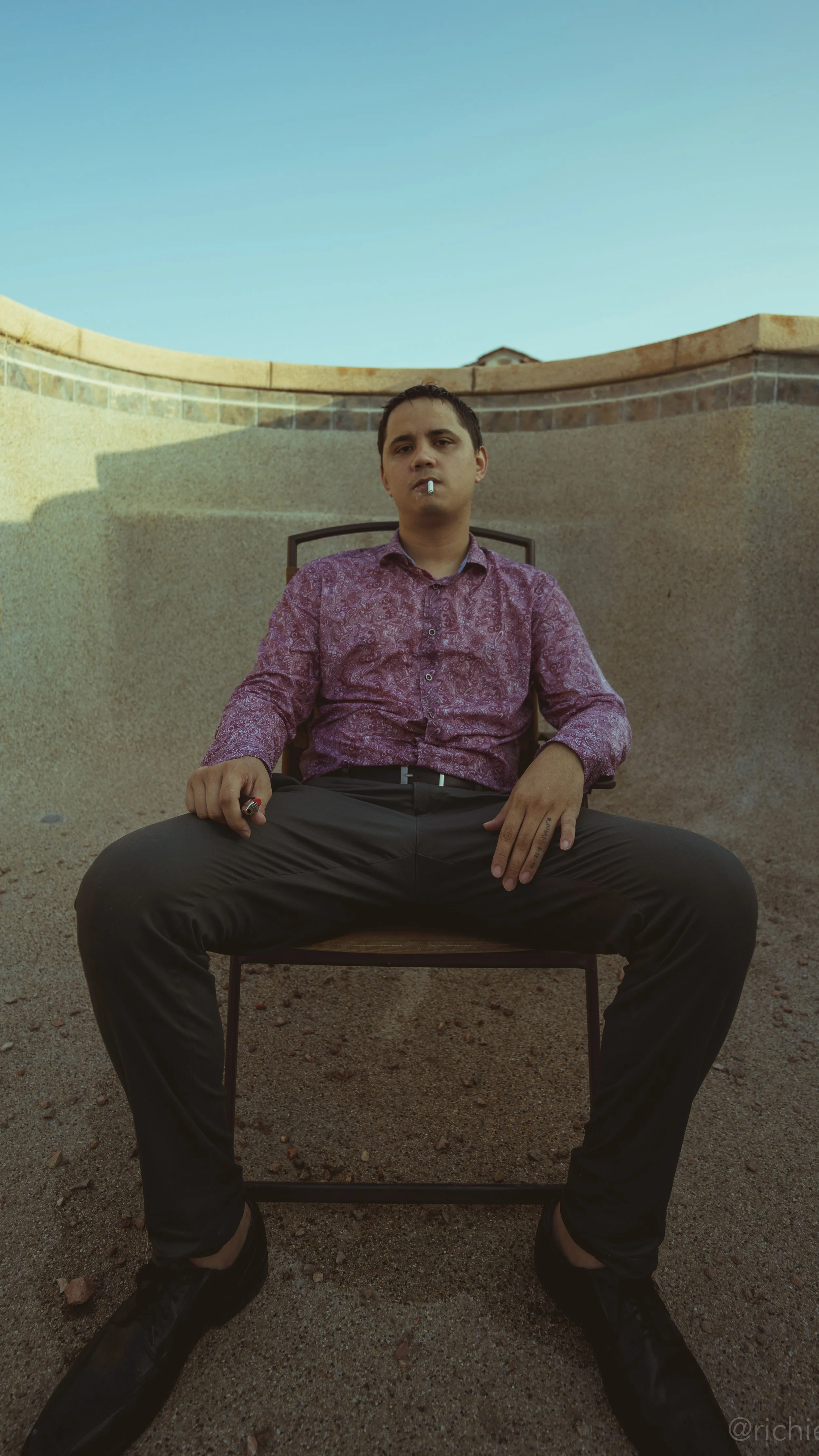 A young man sitting in a chair outdoors on a gravel surface, wearing a purple patterned shirt, black pants, and black shoes, with a cigarette in his mouth and holding a lighter in his hand, with a brick and concrete wall behind him and a clear blue sky above.