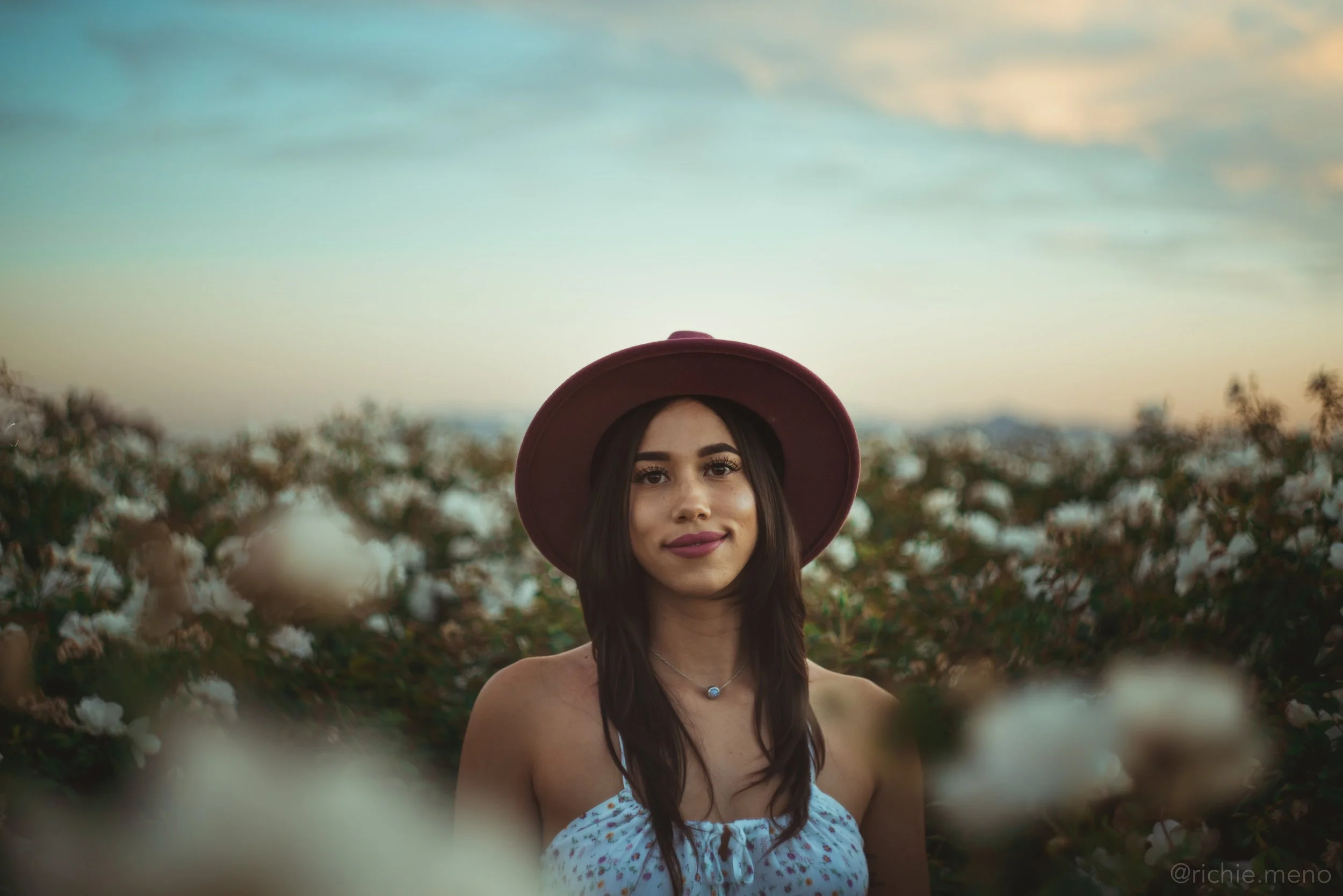 A young woman with long dark hair wearing a maroon hat and a white strapless top, standing in a field of blooming white flowers at sunset.