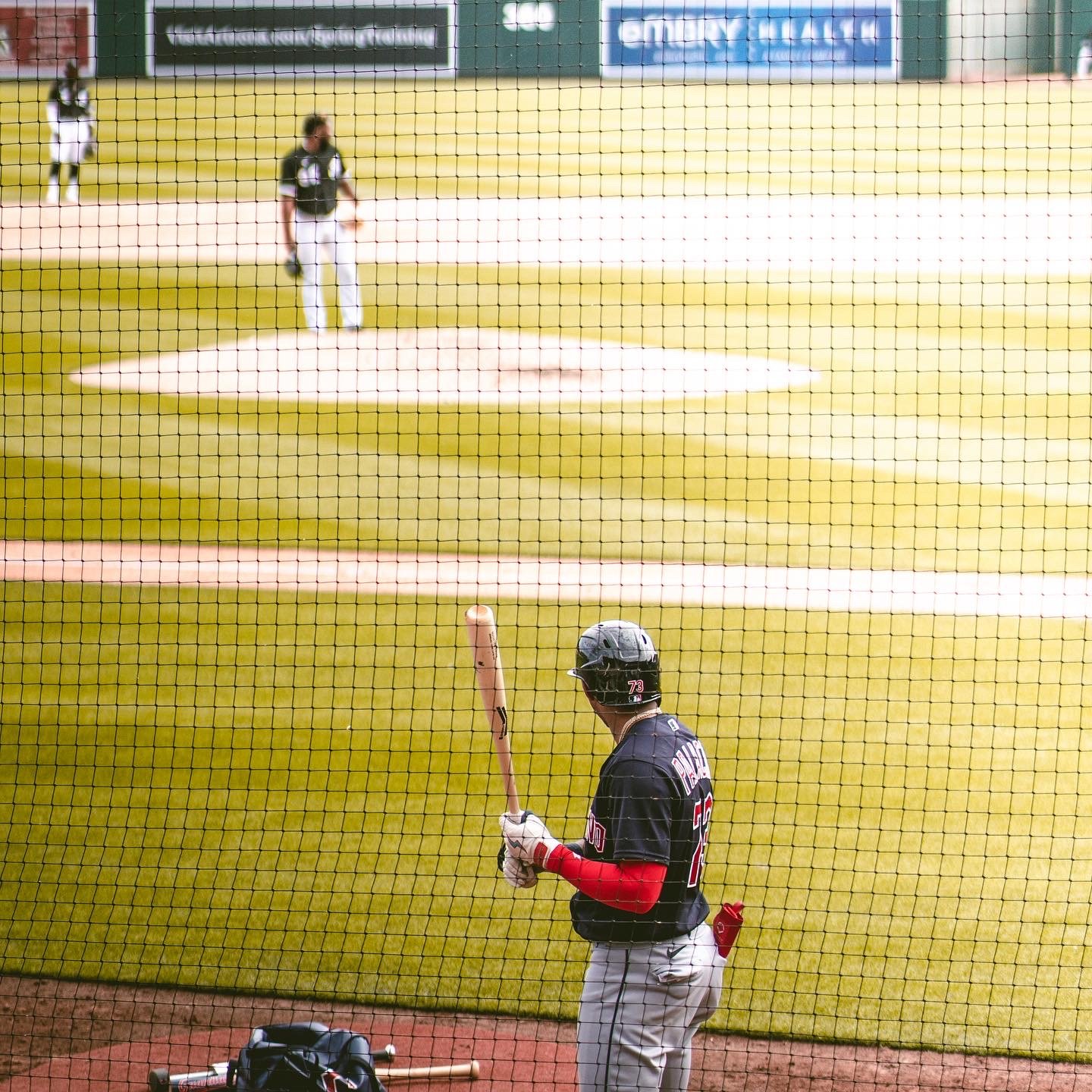 Baseball player standing in the dugout holding a bat, viewed through a protective fence, looking towards the field where other players are practicing.