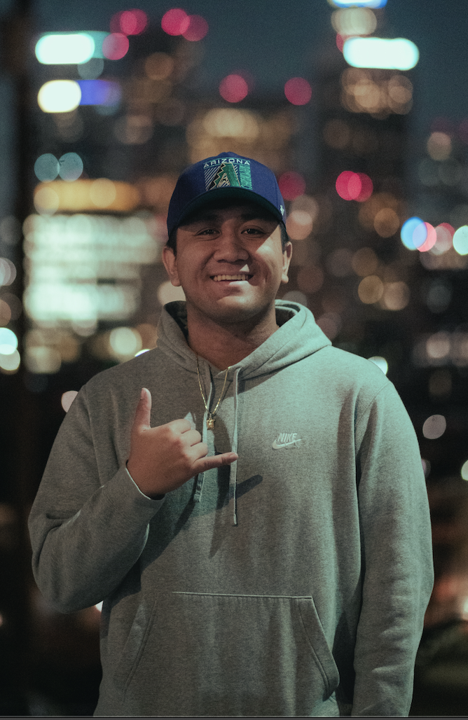 Young man smiling outdoors at night, wearing a gray Nike hoodie and a blue cap with roadside scenery blurred in the background.