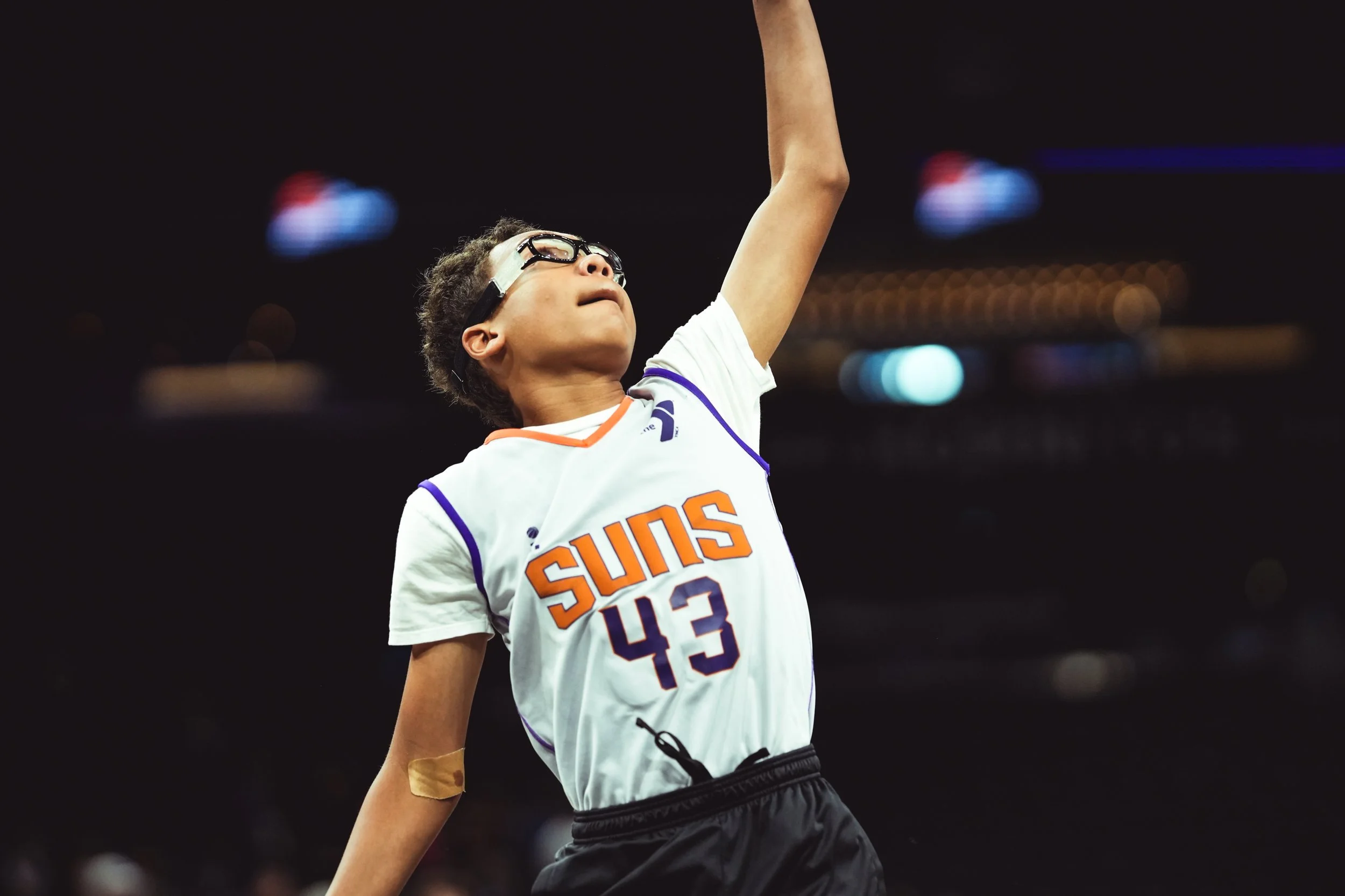 A young boy wearing glasses and a Phoenix Suns basketball jersey with the number 43, making a basketball shot.