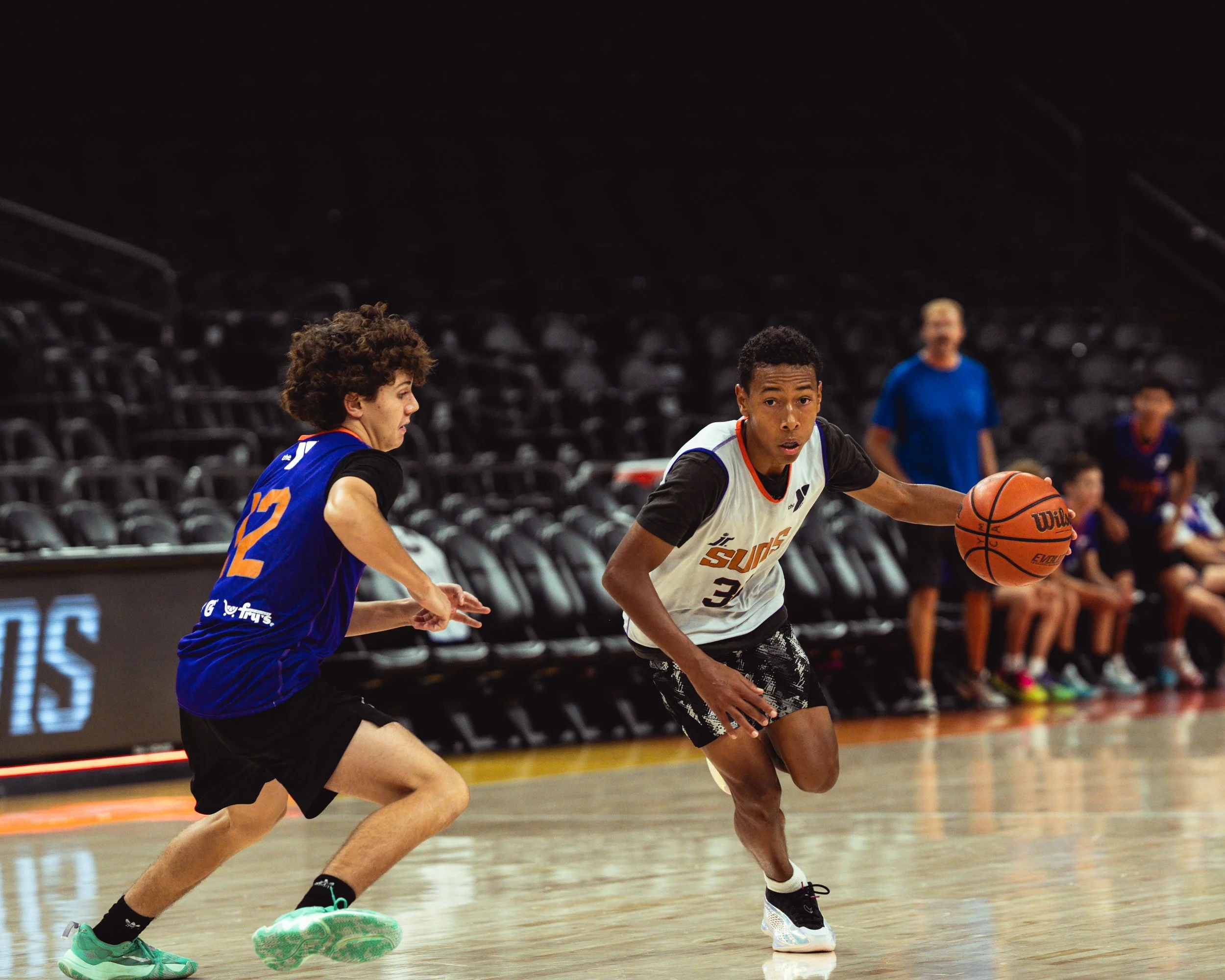 Two young male basketball players in action during a game; one dribbling the ball, the other defending.