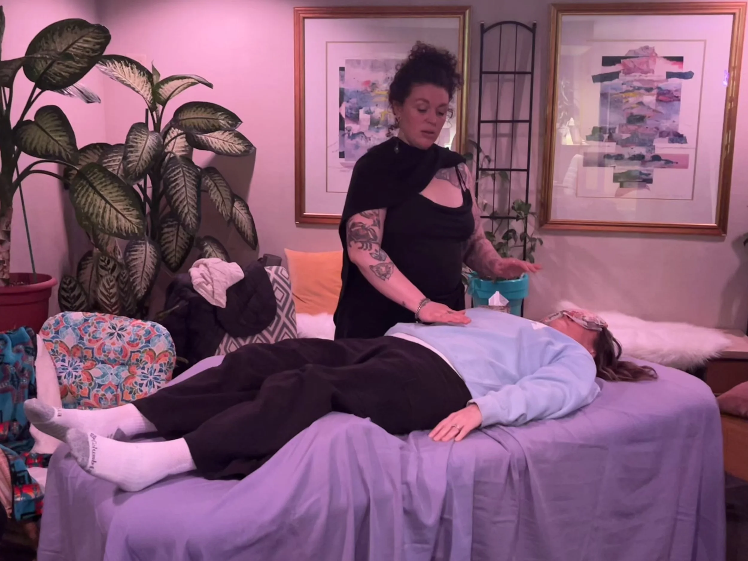 A woman with tattoos on her arms and curly hair styled in an updo, wearing a black top with lace details, is standing beside a bed and appears to be organizing or doing something with her hands. There are multiple people lying on the bed, covered with blankets. The room has purple lighting, framed abstract artwork on the walls, a large potted plant to the left, and a decorative iron window grill in the background.