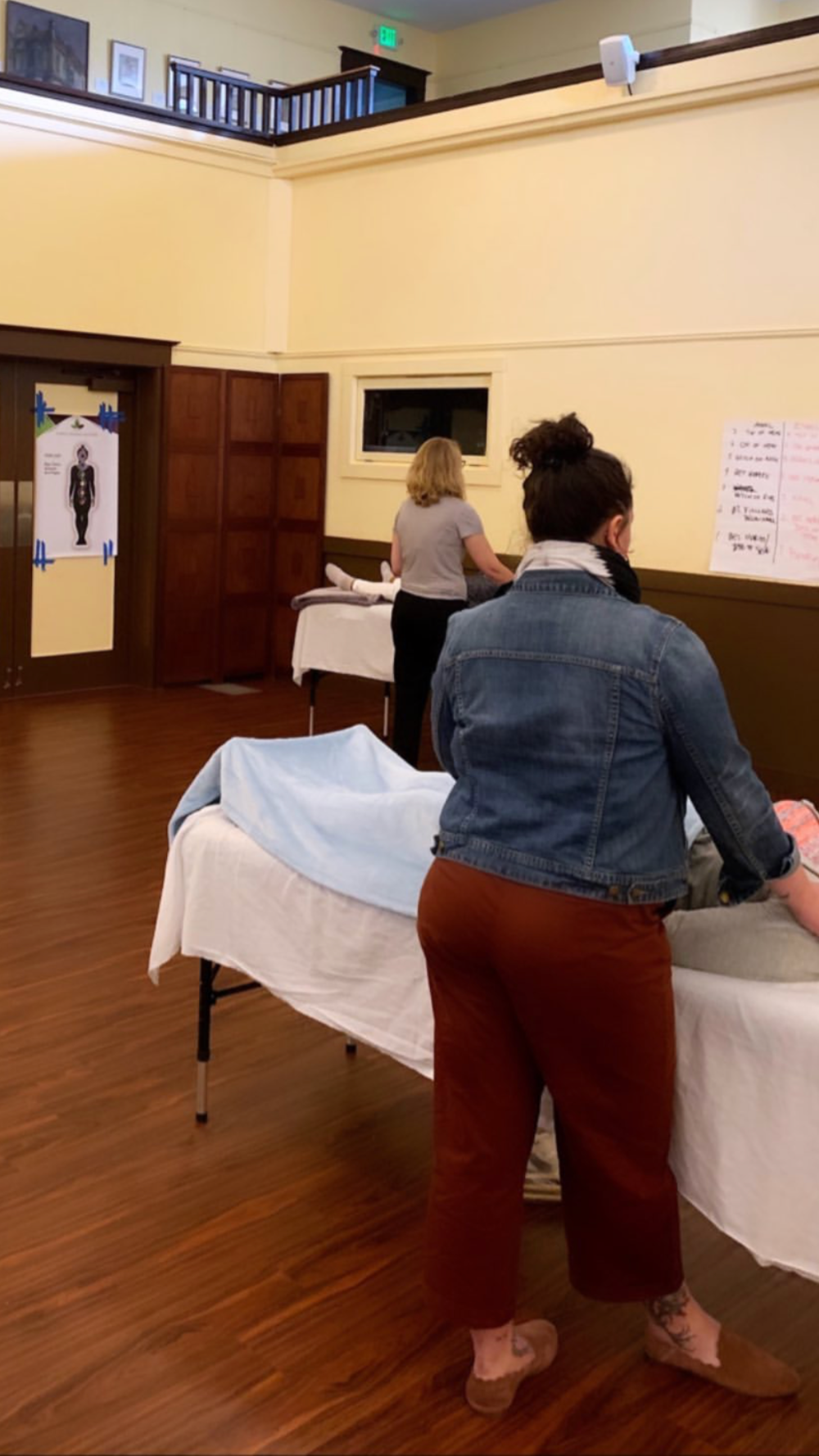 People in a room with massage tables, one person is lying face down while others are standing nearby, and a large diagram of a human figure on the wall.