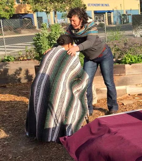 Two women engaging in a prayer or moment of reflection, one kneeling and wrapped in a colorful blanket, and the other standing with arms outstretched, outdoors in a garden with raised beds and a building in the background.