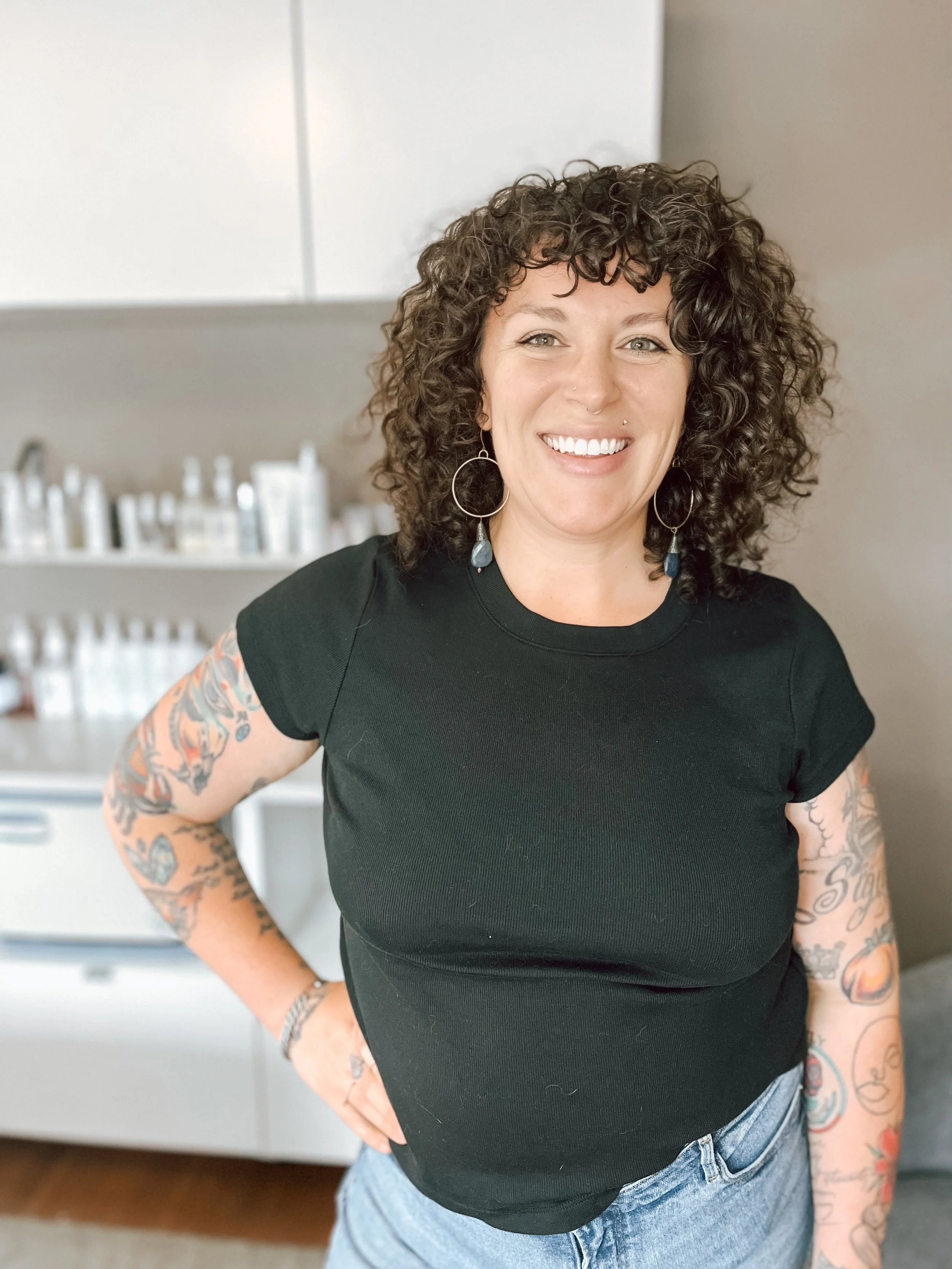 A woman with curly dark hair, tattoos on her arms, wearing a black t-shirt and blue jeans, smiling in a room with shelves of bottles or containers in the background.