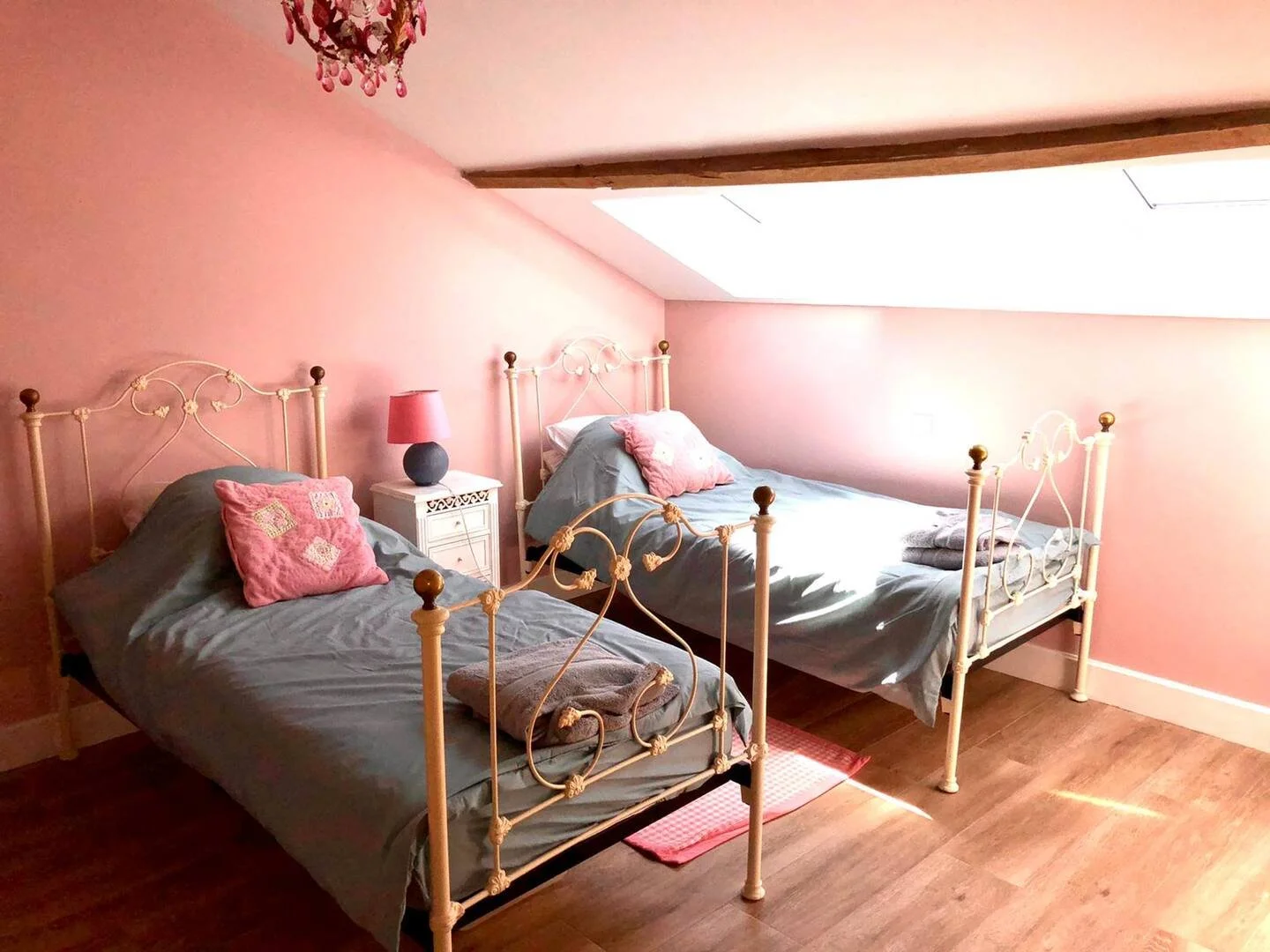 A bedroom with pink walls and two white metal beds, each with a pink pillow and gray bedsheets. Between the beds is a white nightstand with a round pink lampshade, and a towel on one of the beds. There is a skylight window on the sloped ceiling, and