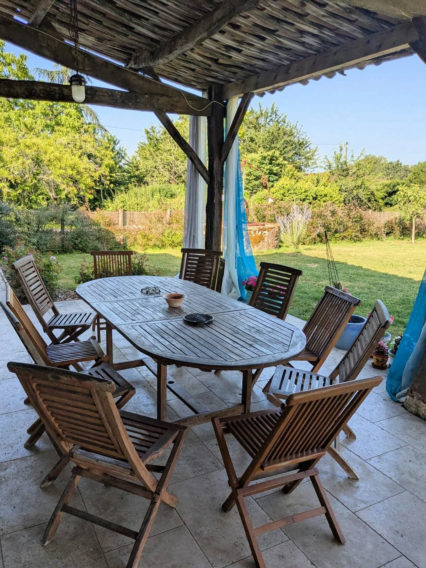 Outdoor patio with a wooden dining table and eight matching wooden chairs, overlooking a green backyard with trees and a wooden fence, shaded by a rustic wooden pergola with hanging lights and blue curtains.