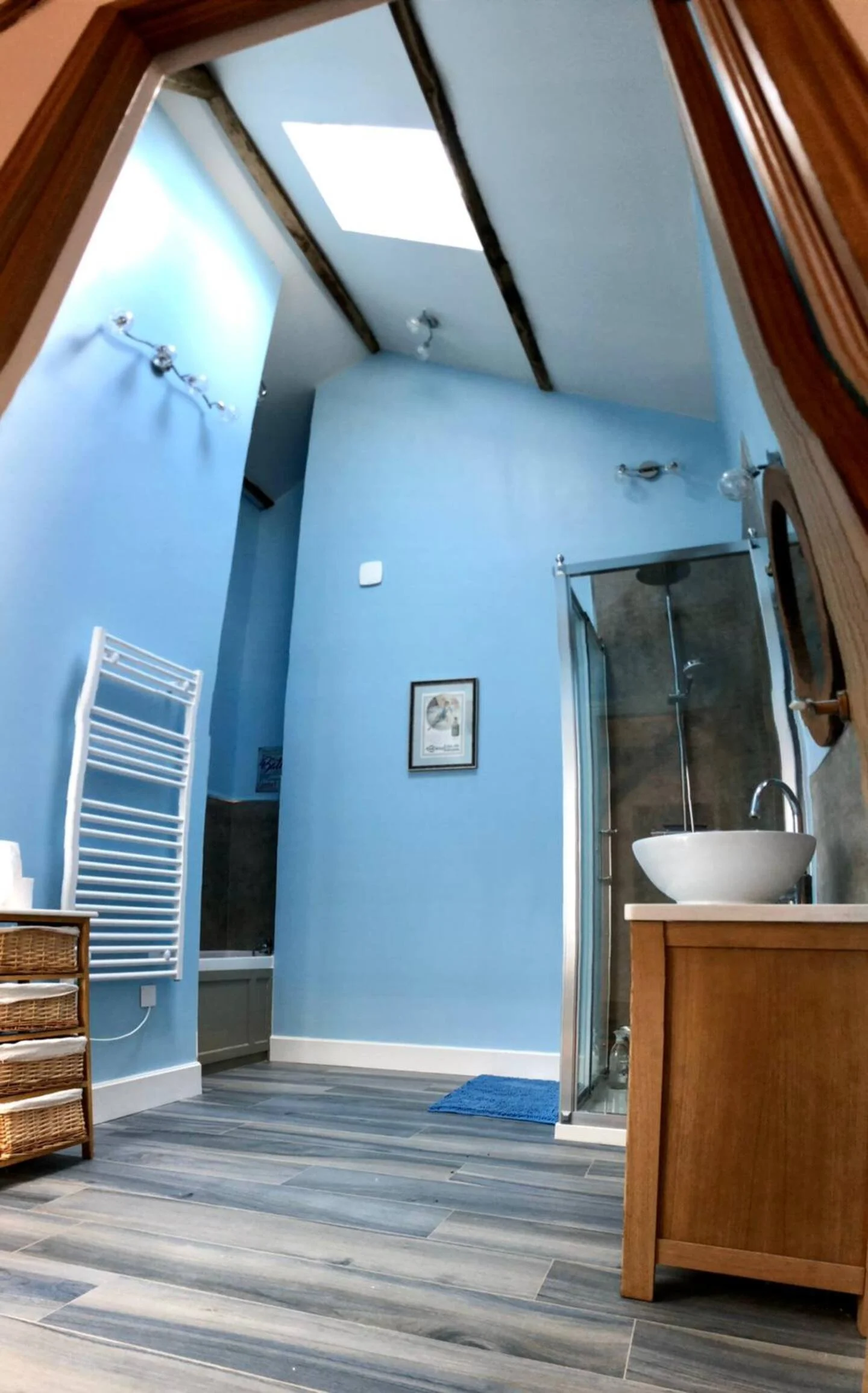 Bathroom with blue walls, skylight, wall-mounted lights, wicker basket storage, framed picture, towel radiator, shower enclosure, sink on wooden vanity, gray wood-look floor, ceiling beams.