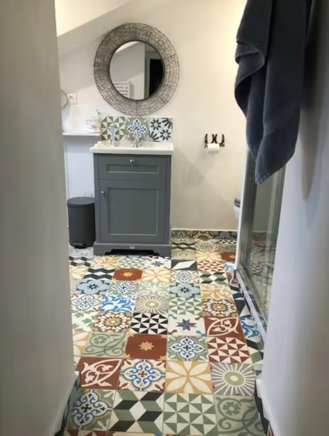 A small bathroom with a colorful patchwork tile floor, a single gray vanity with a decorative mirror above, and a shower enclosure on the right. There is a black trash can next to the vanity and a towel hanging on the right side.