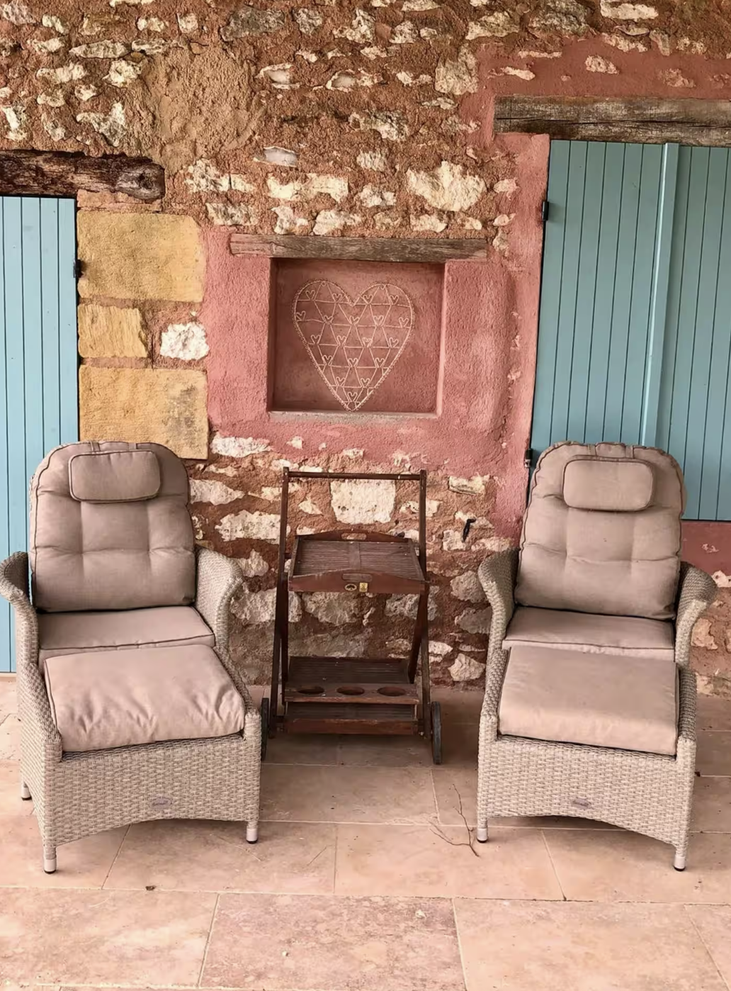 Two cushioned beige armchairs with wicker bases are placed side by side, with a small, vintage wooden bar cart between them, in front of a rustic stone and pink plaster wall. Above the chairs, there's a small recessed wall niche with a heart-shaped w