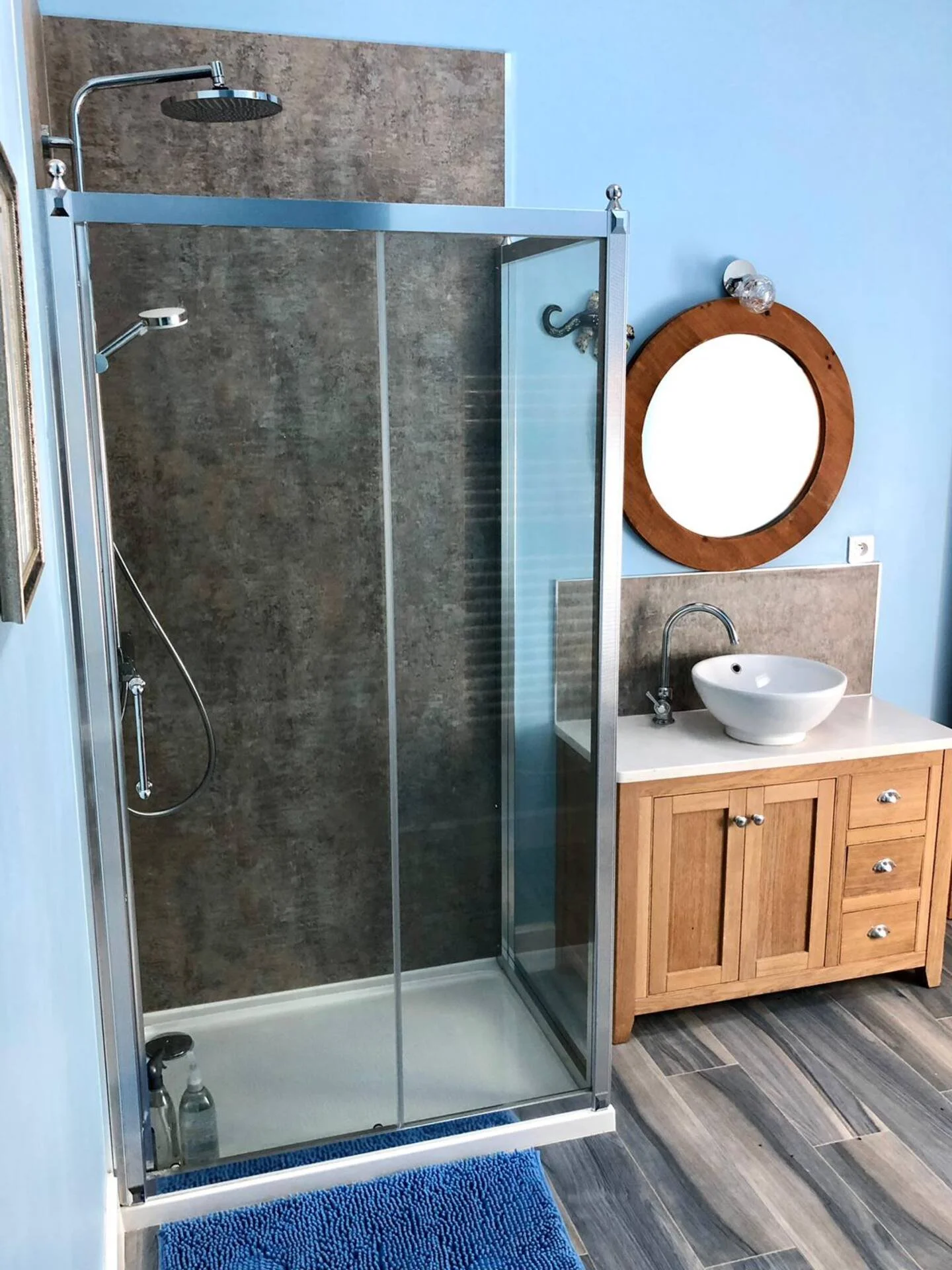 A bathroom with a glass shower enclosure, a wooden vanity with a vessel sink, a round mirror, and a blue bath mat on a striped wooden floor.