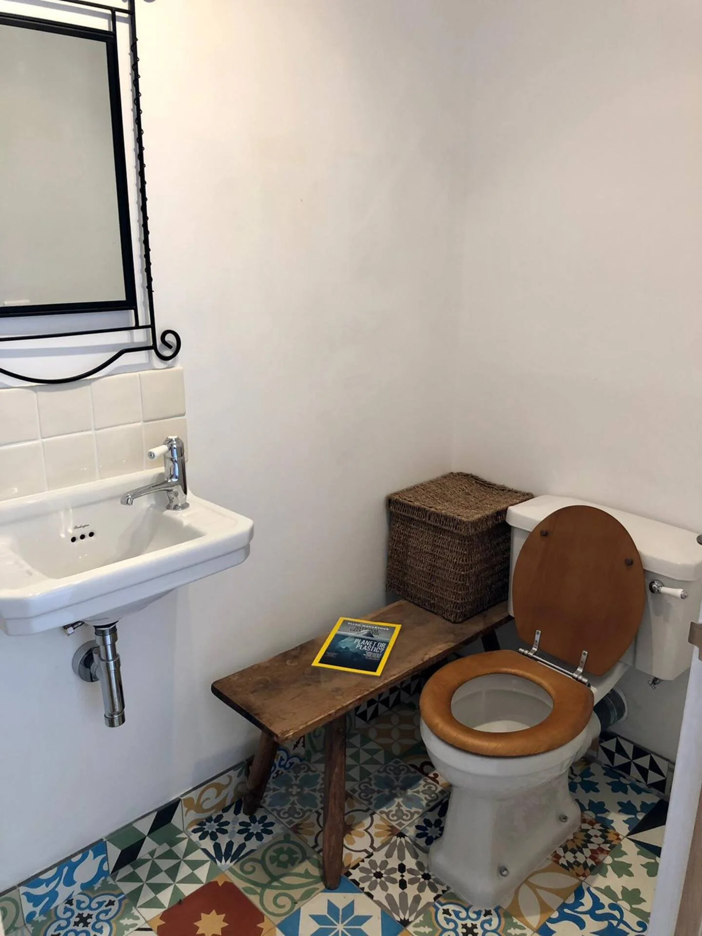 A small bathroom featuring a vintage toilet with a wooden seat and lid, a wooden bench with a magazine and a decorative basket on top, a white porcelain sink with a chrome fixture, a small mirror with a black metal frame, and colorful patterned tile