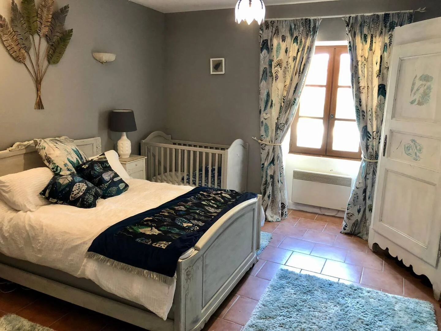 A cozy bedroom with a white wooden bed, nautical-themed pillows, a dark bedside lamp, a white crib, patterned curtains, a window with sunlight, a white wardrobe, and a fluffy rugs on terracotta-tiled floor.
