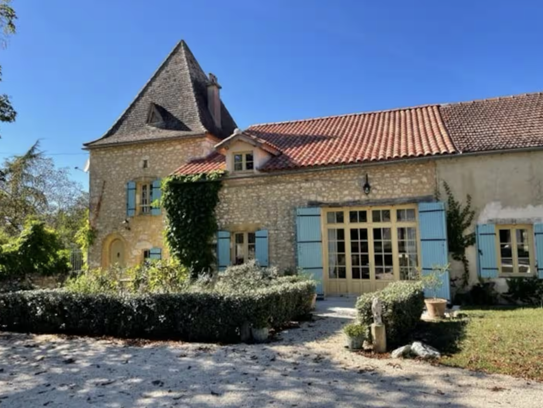 A charming stone house with a steeply pitched roof, blue shutters, and a garden with bushes and potted plants under a clear blue sky.