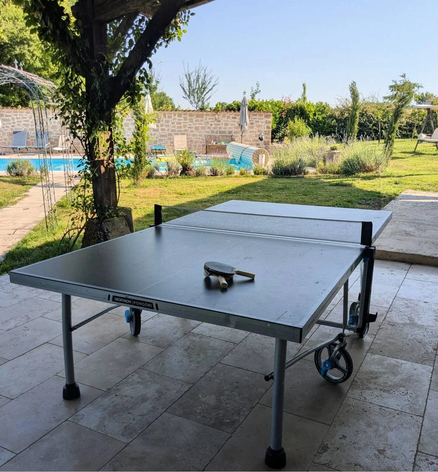 Outdoor ping pong table with a paddle and ball on top, set up under a shaded patio with a garden, swimming pool, and lounge chairs in the background.