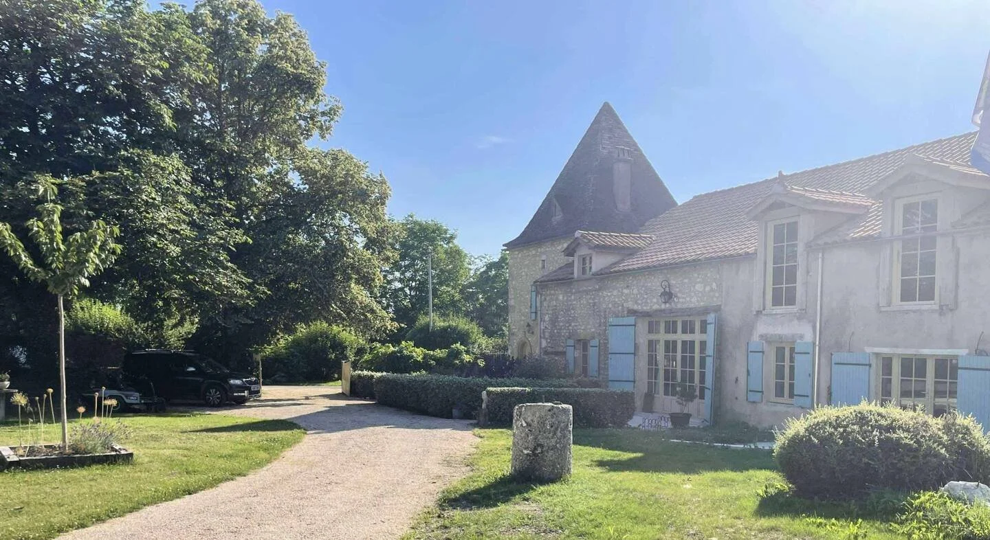 A stone house with blue shutters and a conical tower, surrounded by green trees and bushes, on a sunny day with clear blue sky.