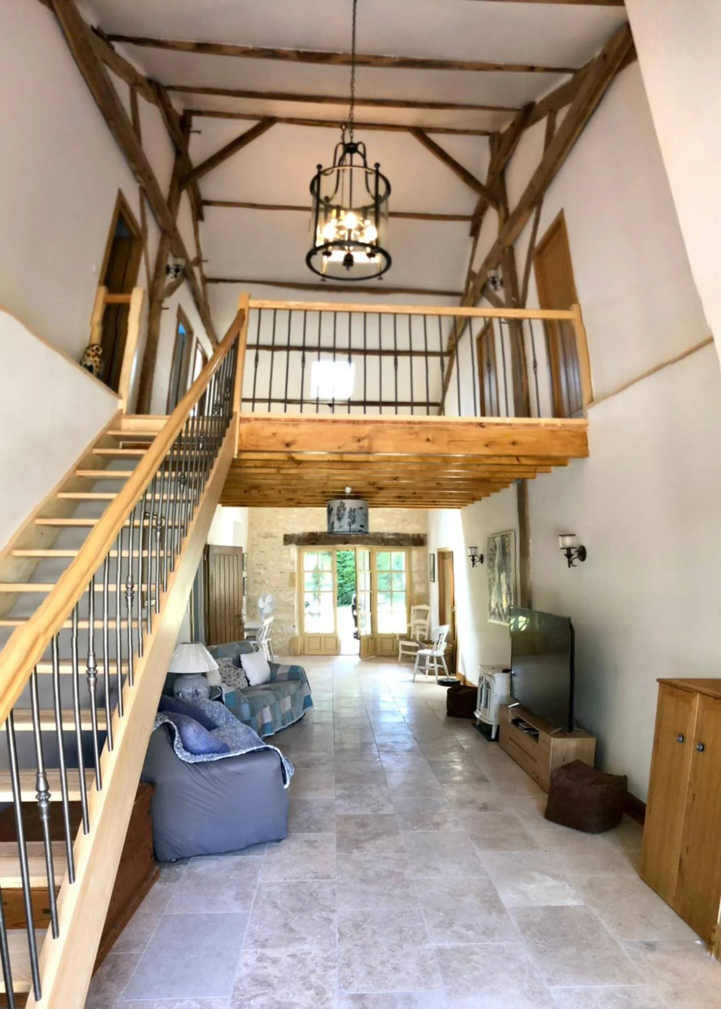 A spacious living room with beige tiled floor, a couch, chairs, a TV, and wooden accents on the walls and ceiling. There is a staircase leading to a loft area with a chandelier hanging from the ceiling.