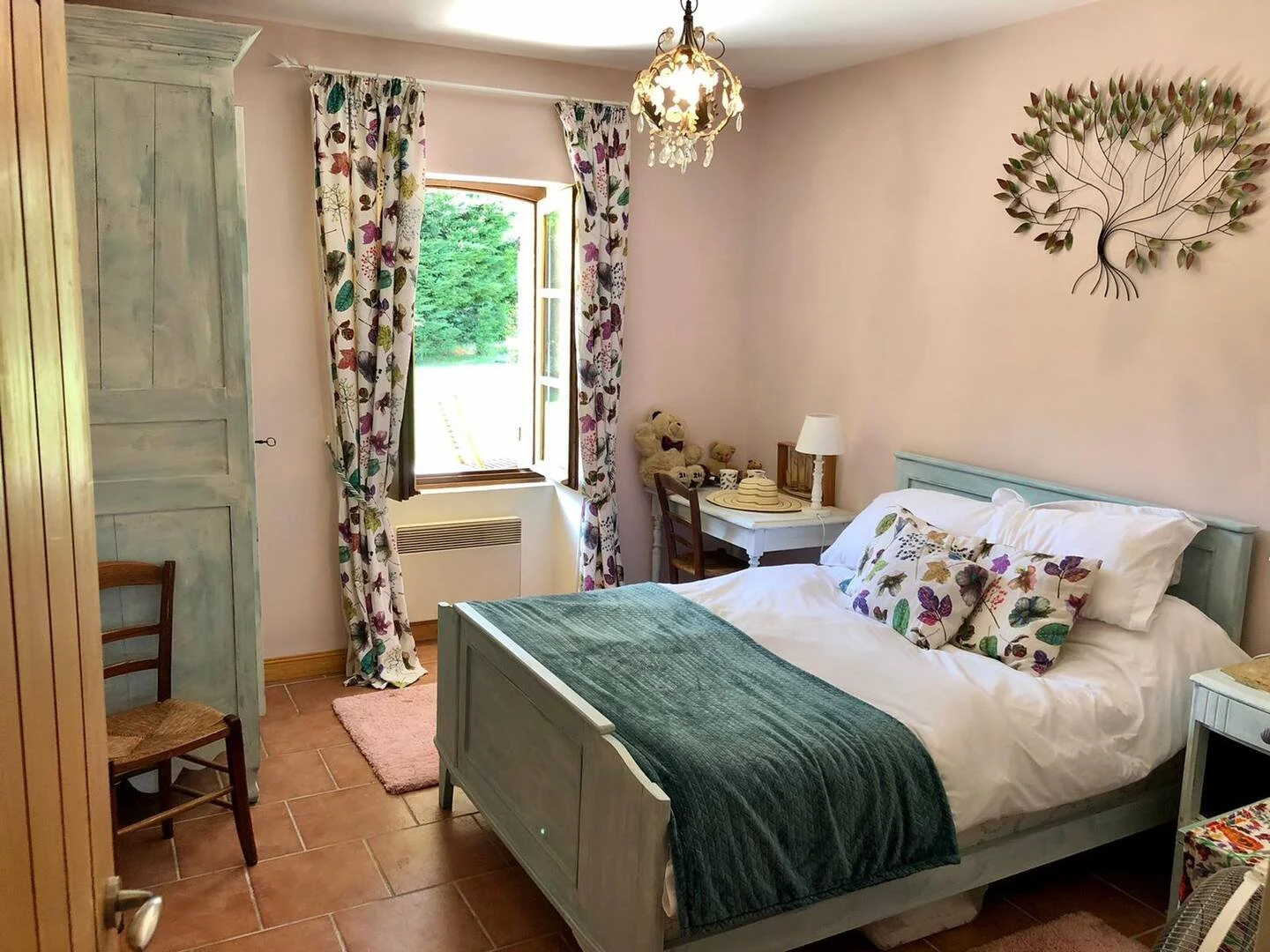 Cozy bedroom with a white bed topped with butterfly-patterned pillows and a teal blanket, a window with floral curtains open to green outdoors, a vintage dresser, a small desk with a lamp and teddy bear, and decorative wall art resembling a tree.