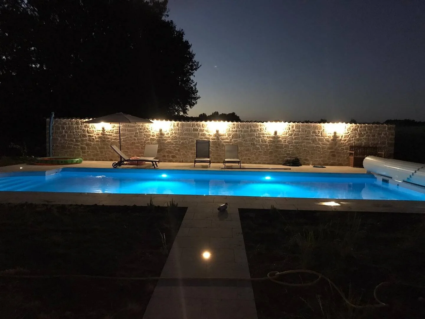 Nighttime view of a backyard swimming pool illuminated with blue lights, surrounded by a stone patio, two lounge chairs with an umbrella, and a stone wall with wall-mounted lights in the background.