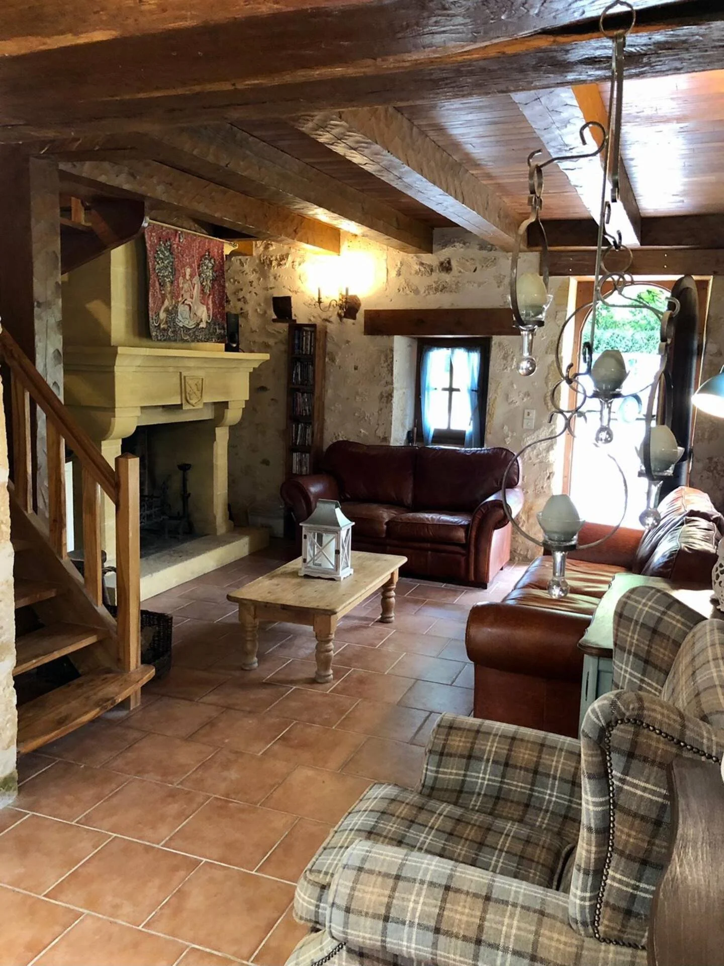 Cozy living room with wood and stone walls, wooden ceiling, leather sofas, plaid armchair, coffee table, decorative chandelier, fireplace, and window with blue curtains.