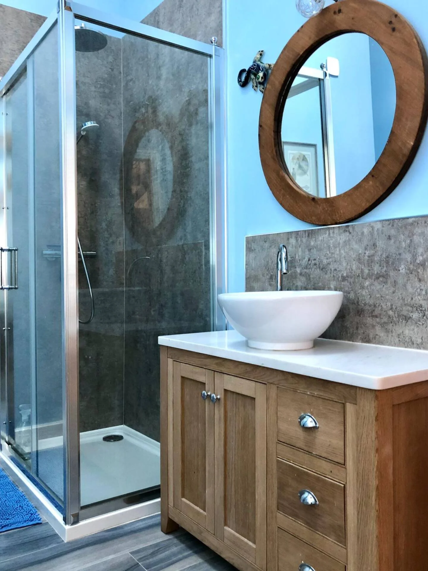 A bathroom featuring a glass shower enclosure with a corner entrance, a white vessel sink on a wooden vanity with three drawers, a round wooden-framed mirror above the sink, a blue wall, and a small decorative object on the wall near the mirror.