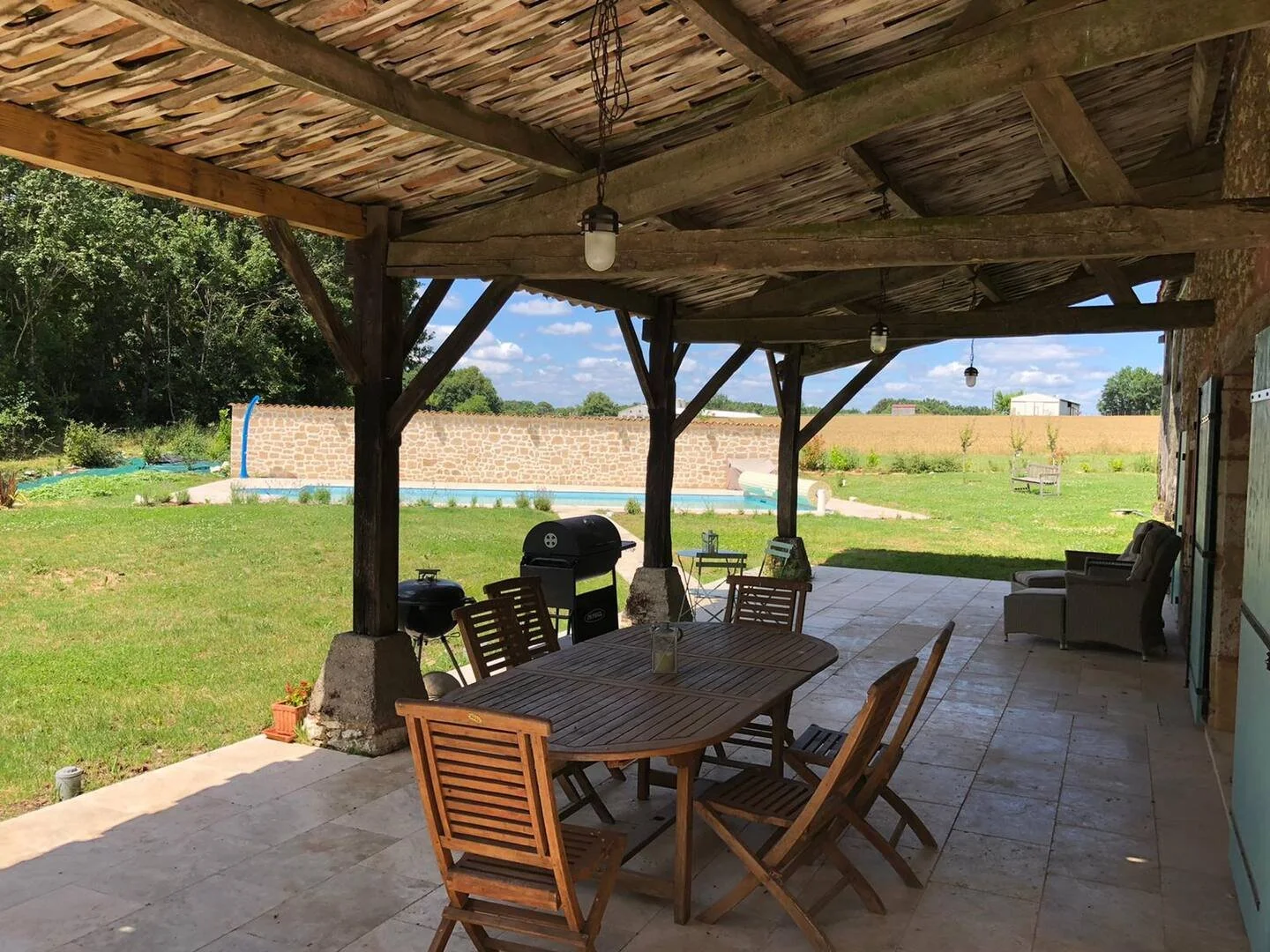 Covered patio with wooden dining table and chairs, outdoor lounge chairs, a barbecue grill, and a swimming pool in a backyard with green grass, trees, and a stone wall, under a bright blue sky.