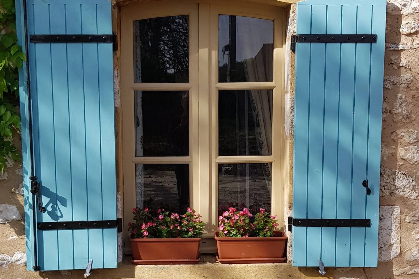 A window with blue wooden shutters and a beige frame, with two flower boxes filled with pink flowers on the window sill, attached to a stone wall.