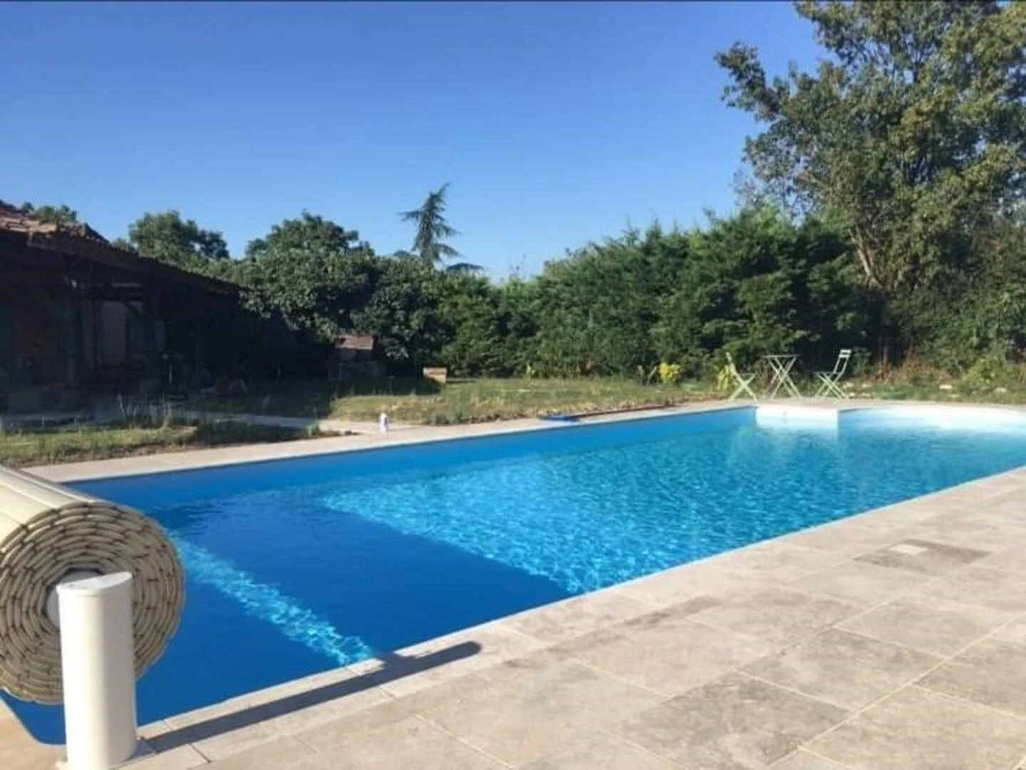An outdoor rectangular swimming pool filled with blue water, surrounded by a gray stone patio, with green trees and bushes in the background and a clear blue sky overhead.