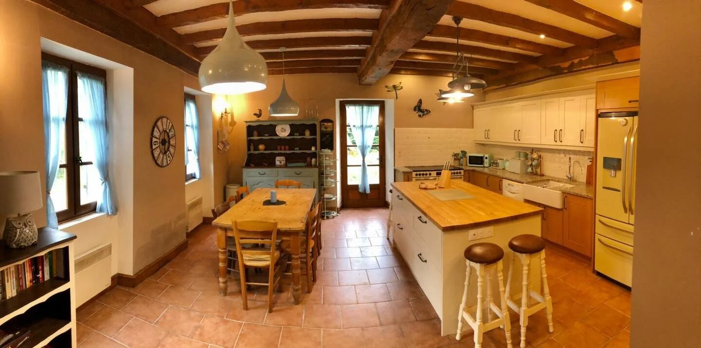 A cozy kitchen and dining area with wooden beams on the ceiling, two windows with blue curtains, a wooden dining table with six chairs, a kitchen island with two barstools, cream-colored cabinets, and a yellow refrigerator.