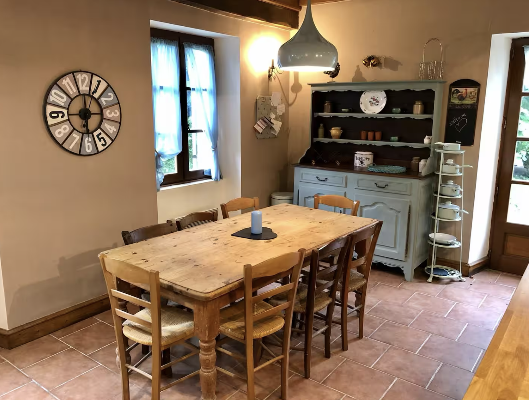 Rustic kitchen dining area with a wooden table and six chairs, a vintage blue hutch with dishware, a large wall clock, and a window with blue curtains letting in natural light.