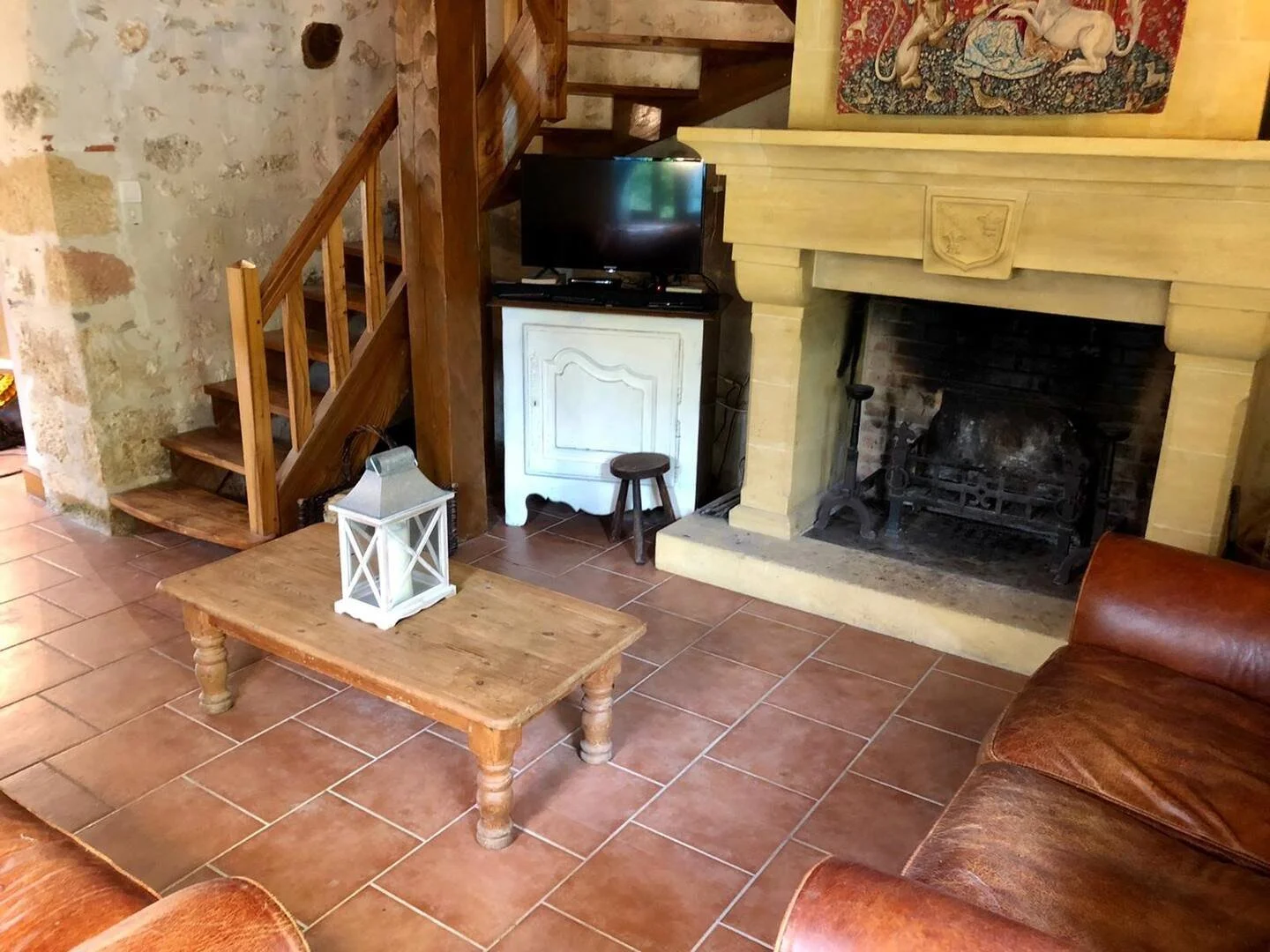 A cozy living room with terracotta tile flooring, a fireplace with a carved stone mantle, a leather sofa, a wooden coffee table with a lantern on top, a small TV on a white cabinet, a rustic wooden staircase, and artwork hanging above the fireplace.