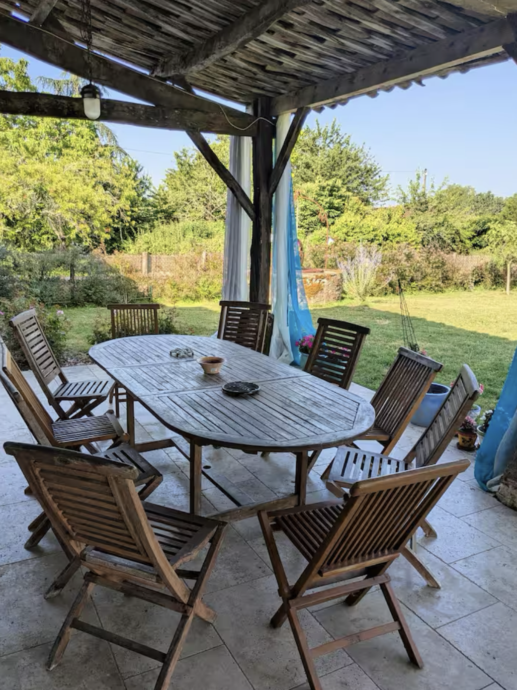 Wooden patio table with multiple chairs under a covered porch, overlooking a garden with trees and shrubs, some potted plants, and a grassy area in the background.