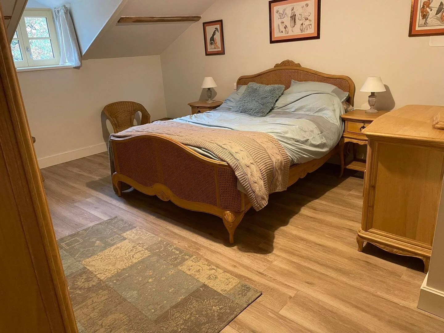 A cozy bedroom with a wooden bed frame, white bedding, and a gray pillow. There are two wooden nightstands with lamps on each side of the bed, a wicker chair in the corner, and framed artwork on the wall. A window with white curtains lets in natural