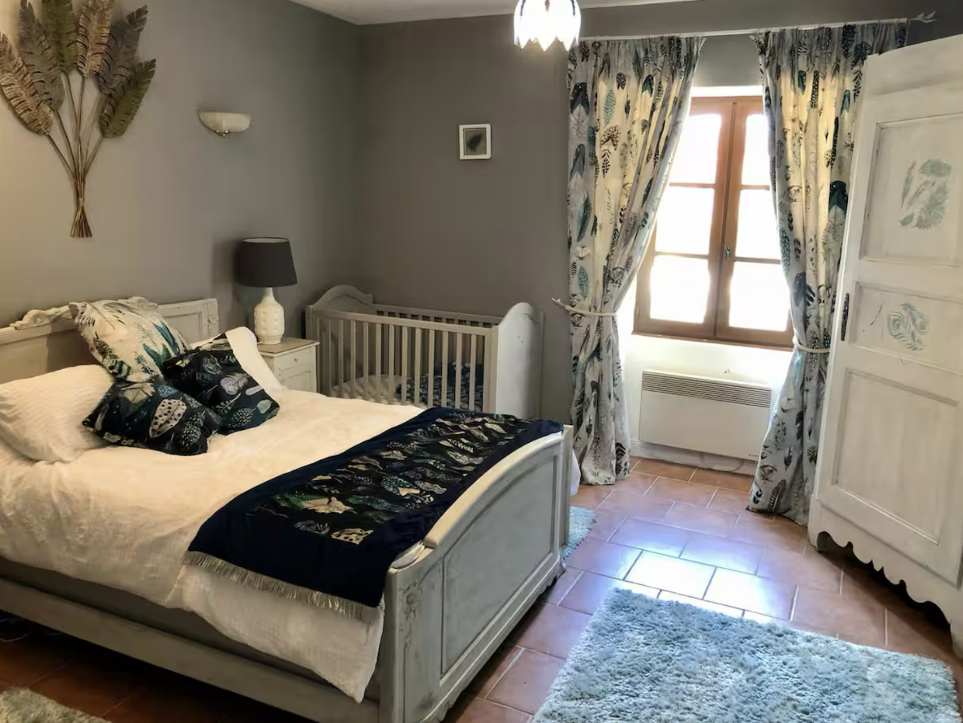 A cozy bedroom with a white bed, blue and white bedding, and pillows. There is a black and white lamp on a white nightstand, a white wardrobe with floral designs, a window with blue patterned curtains, a small white framed picture on the wall, and a