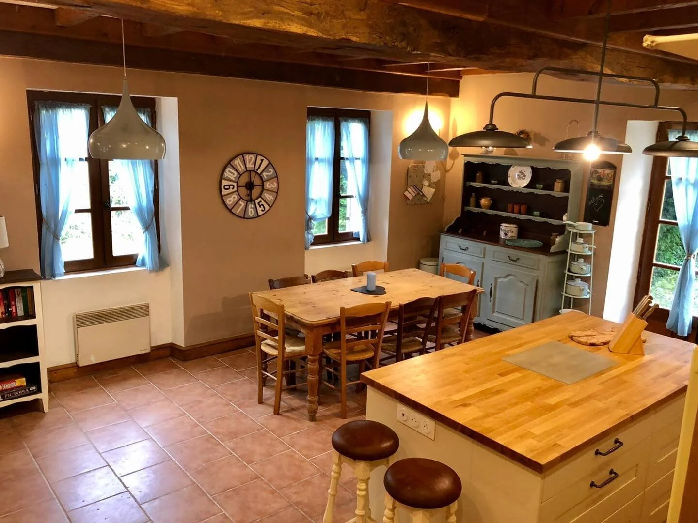 Photo of a rustic kitchen and dining area with wooden beams, tiled floor, a dining table with six chairs, three windows with blue curtains, a wall clock, a light-colored cabinet with dishes, and a kitchen island with two stools.