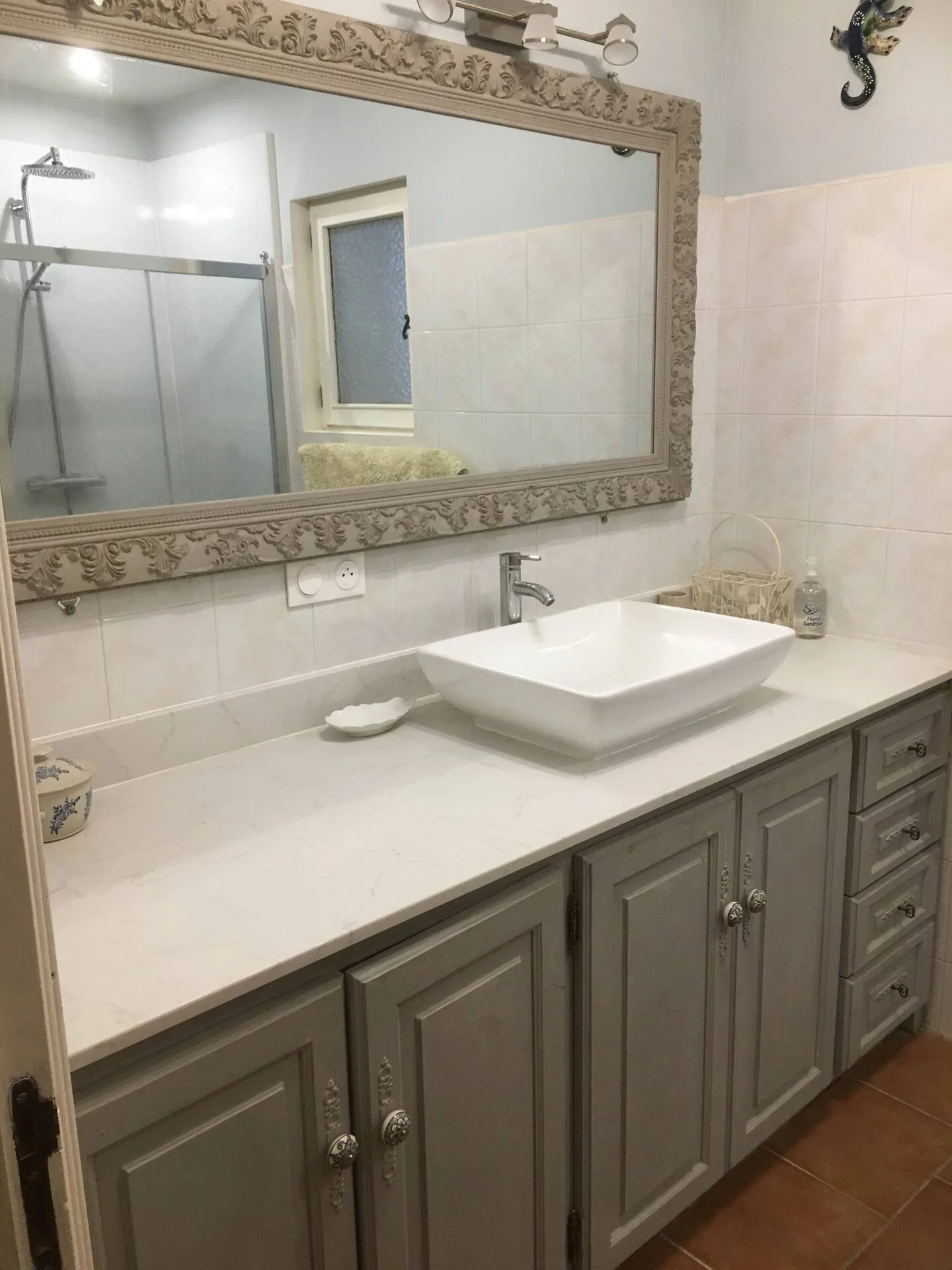 A bathroom sink vanity with a large ornate mirror, a rectangular vessel sink, a faucet, and decorative items including a soap dispenser and a small dish. In the background, there is a shower area with glass doors, a window, and a towel.