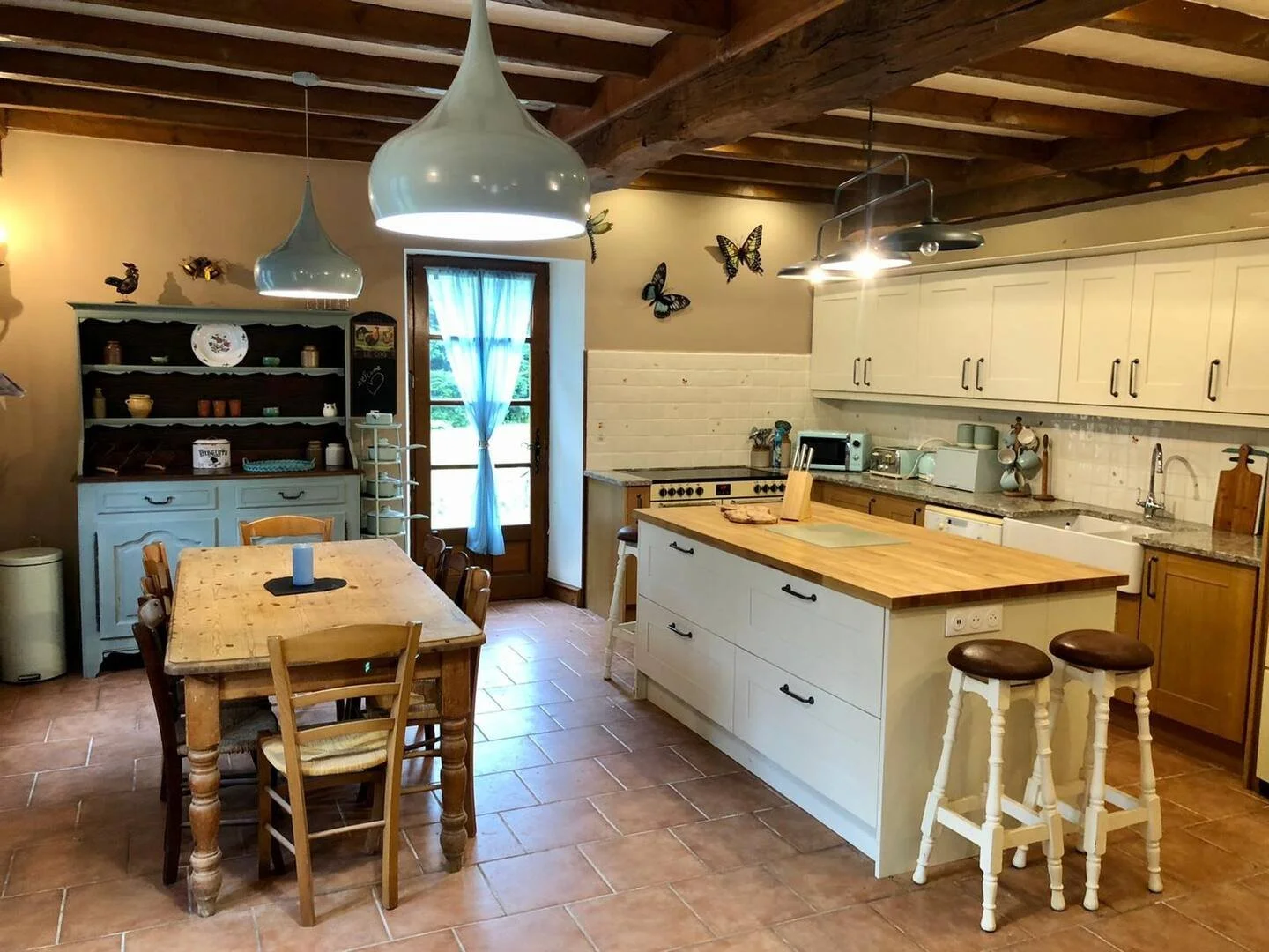 A rustic kitchen with a wooden dining table and chairs, white kitchen cabinets, a kitchen island, and a door with a blue curtain leading outside. Decor includes butterfly wall art, a hutch with dishes, and hanging pendant lights.