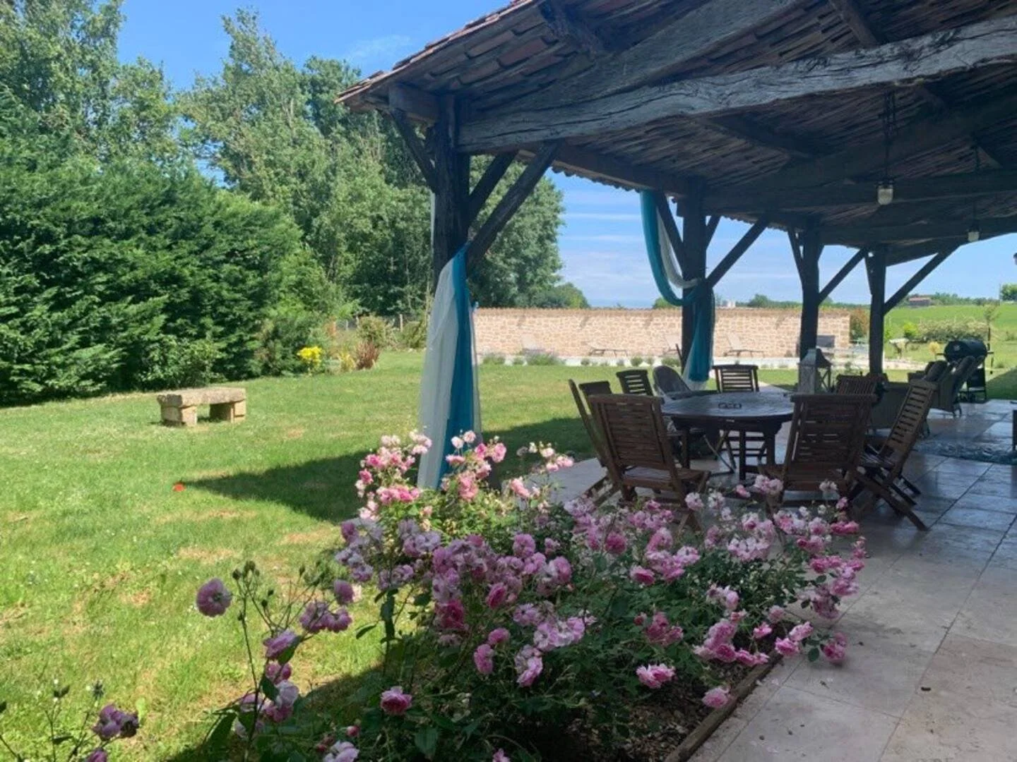 A covered outdoor patio with a wooden roof, patio furniture, pink flowers, green grass, trees, and a brick wall in the background, on a sunny day.